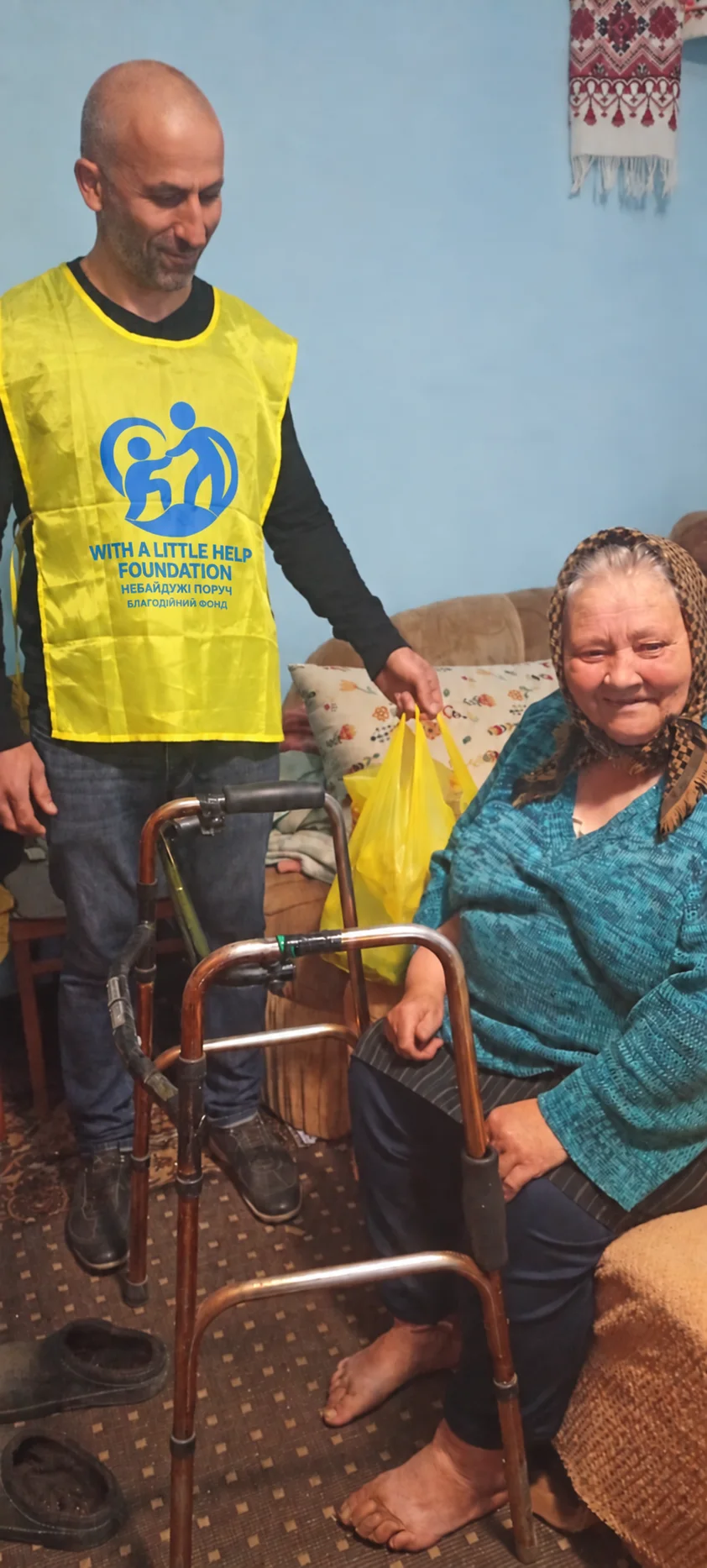 A team member in a yellow vest beside a seated woman and a yellow bag of supplies in her home.